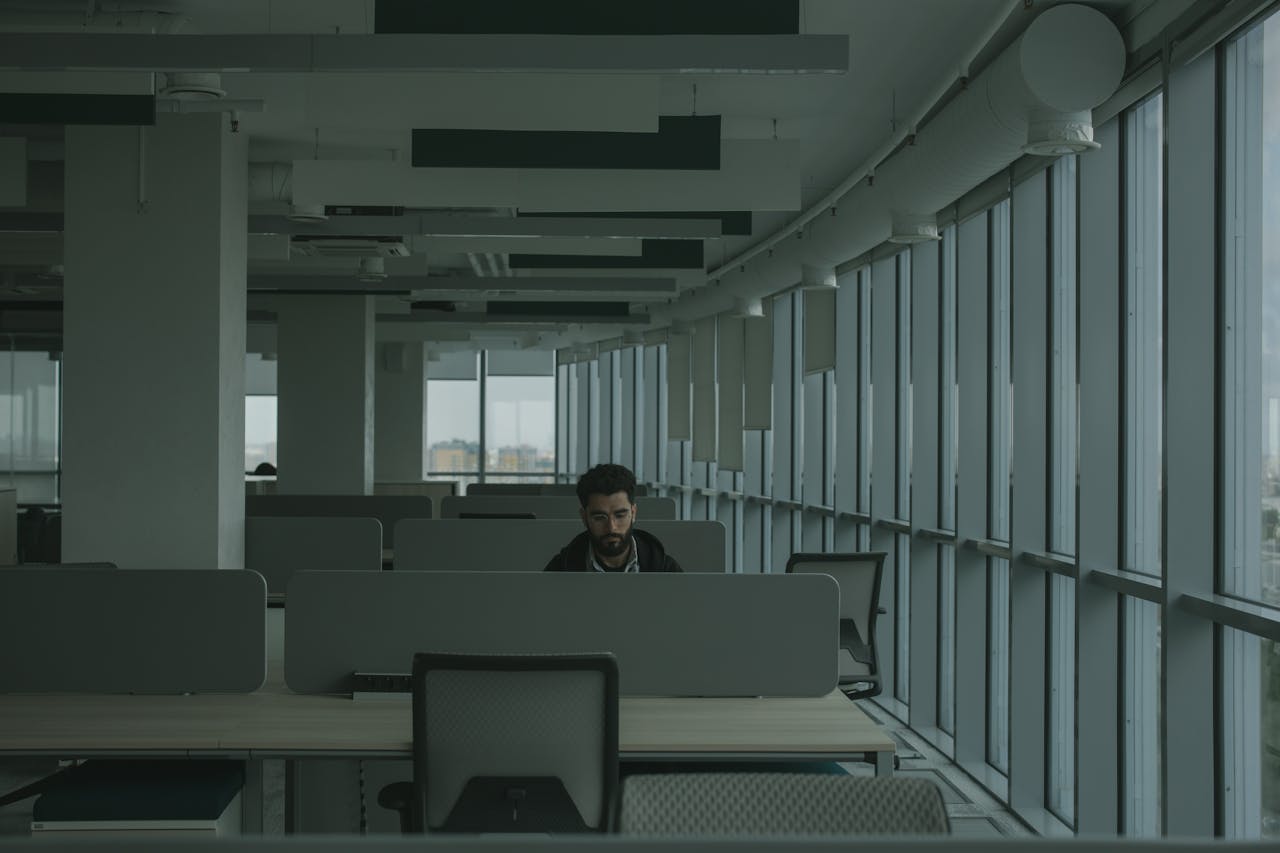 An adult man works alone in a contemporary open-plan office, surrounded by desks and dividers.